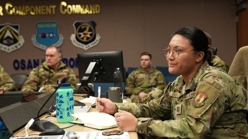 Multiple airmen are present in the image but one sits in the forefront, all listening to a brief within a conference room.