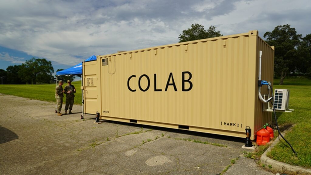 A large cargo container pained desert brown has the word COLAB on the side in large lettering. Two airmen stand next to the entrance of the cargo container.