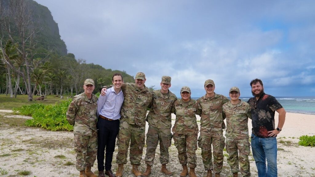 Group of airmen and civilians stand together on a beach together, they are all facing towards the camera, smiling.