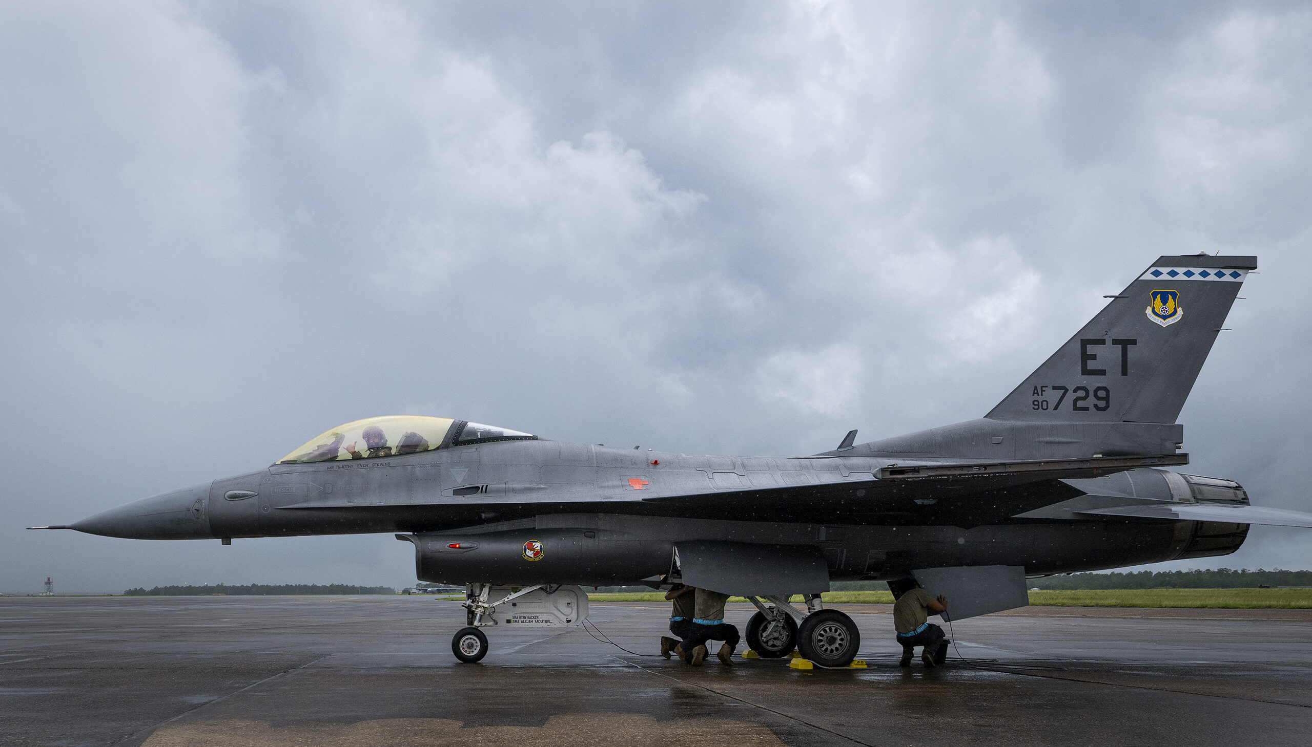 Profile view of full F-16 in the rain. The pilot is giving a thumbs up from the cockpit while a maintainer finishes up work the rear of the aircraft.