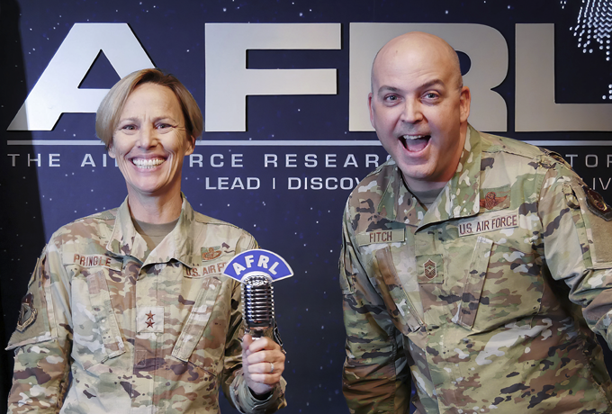 Major General Pringle standing on the left in uniform and smiling isholding a silver microphone with an attachment that reads AFRL. Standing to her right is Chief Master Sergeant Fitch, smiling and in uniform.