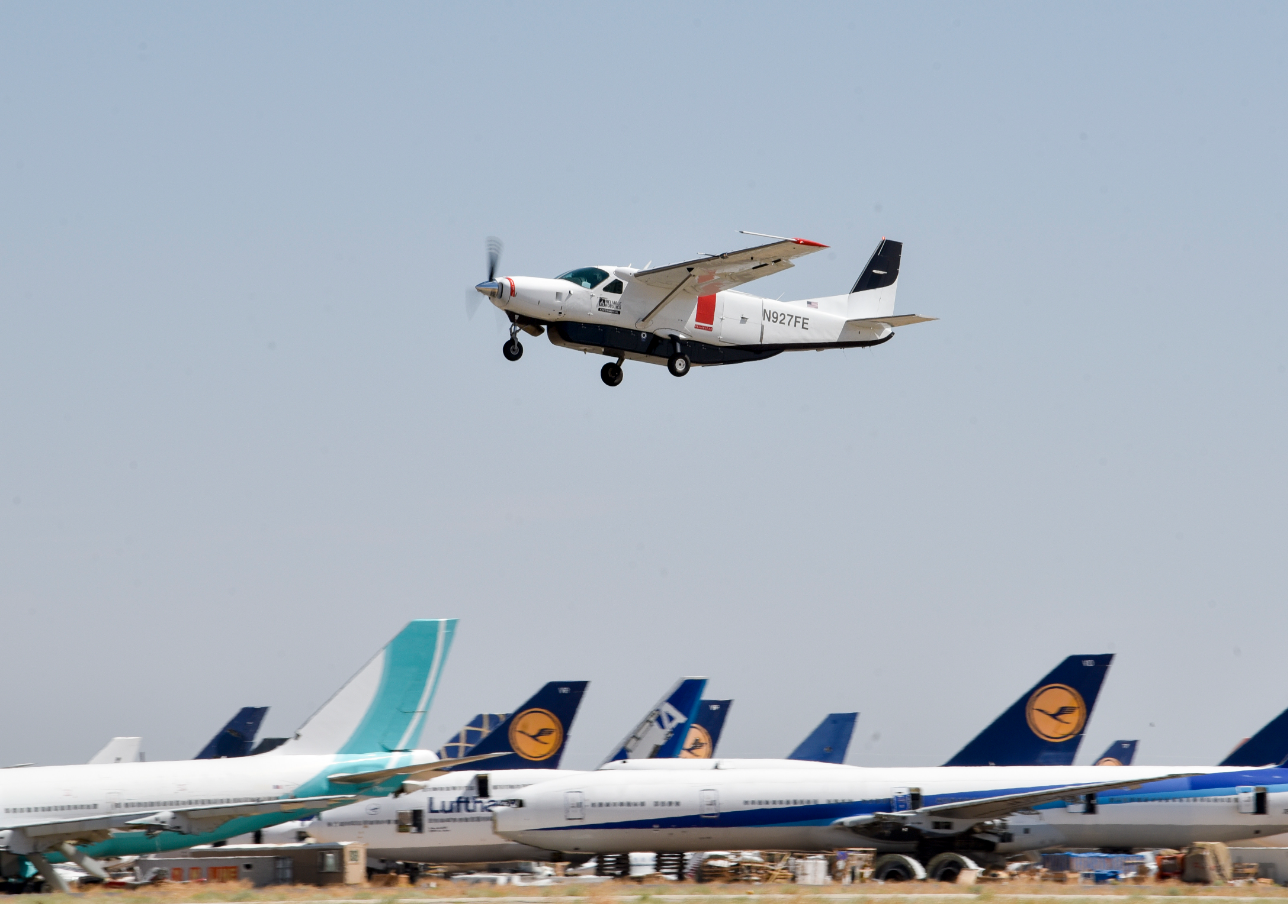 Cessna test craft flies over parked aircraft in open field.