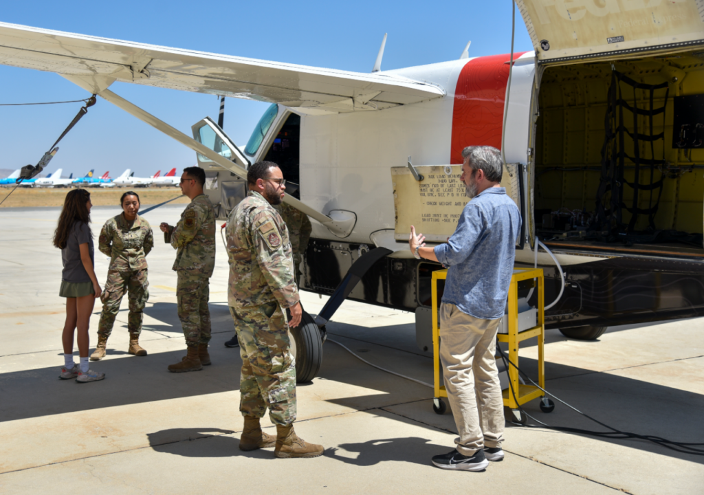 Airmen stand next to a test craft while speaking with another plain clothes team member and a younger individual.