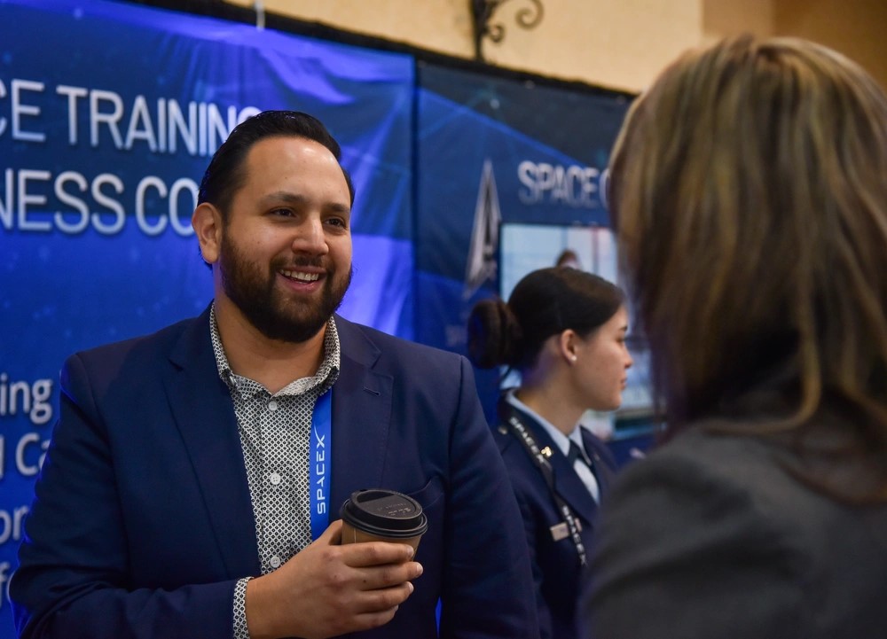 Dr. Arthur Grijalva, Director of SpaceWERX, stands on the left side of the image, speaking to a woman who's facing away from the camera, towards him.